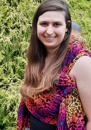 Sunset wreath long woven wrap, size 5, with a demo doll in a ruck back carry with a Tied Tibetan waist-less finish. The wrap is bright purple, yellow, and orange with black ovals arranged in a wreath pattern