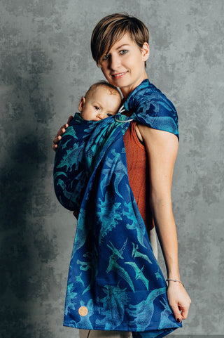 Woman holding a baby in a blue woven ring sling - limited edition Jurassic Park Evolution -against a gray background