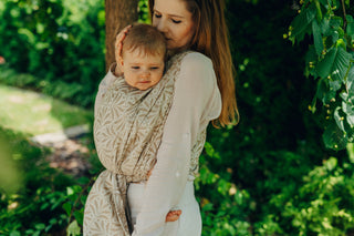 Mother kisses her baby while babywearing outdoors in greenery while wearing her baby in long woven baby wrap in luxury design fabric Infinity Golden Hour, a cotton and bamboo viscose blend woven fabric design.