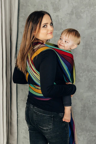 Lenny Lamb Long Woven Wrap - Carousel of Colors. Front carry, young toddler, back view. Wrap has a rainbow of colors in a striped pattern.