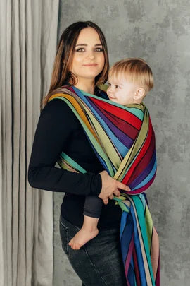 Lenny Lamb Long Woven Wrap - Carousel of Colors. Front carry, young toddler, side view. Wrap has a rainbow of colors in a striped pattern.