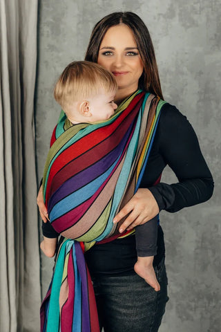 Lenny Lamb Long Woven Wrap - Carousel of Colors. Front carry, young toddler, front-side view. Wrap has a rainbow of colors in a striped pattern.