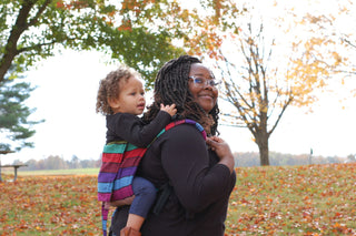 Black woman carrying mixed toddler on her back in Carousel of Colors onbuhimo. Fall trees are in the background