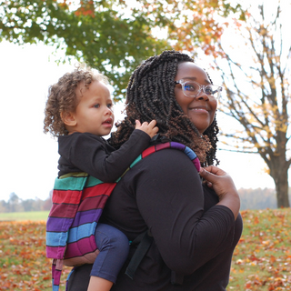 A Black mother carries her biracial baby in a Carousel of Colors onbuhimo in a high back carry. The child is smiling and playing with her mother's hair. Mom is smiling. Side view, outdoors with fall trees behind, light a halo behind the mother's head, and fall leaves on the grass in the backdrop.