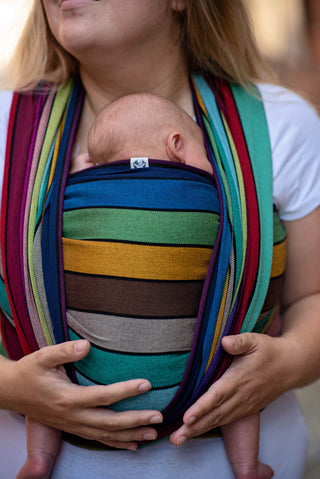 Lenny Lamb Long Woven Wrap - Carousel of Colors - newborn in front wrap cross carry.  Wrap has horizontal stripes in rainbow colors with thin black stripes in-between the colors