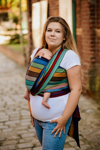 Lenny Lamb Long Woven Wrap - Carousel of Colors - newborn front wrap cross carry side view. Wrap has horizontal stripes in rainbow colors with thin black stripes in-between the colors