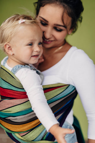 Lenny Lamb Long Woven Wrap - Carousel of Colors - toddler in torso carry front. Wrap has horizontal stripes in rainbow colors with thin black stripes in-between the colors