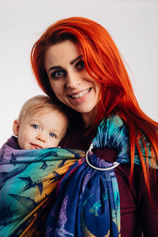 Model wearing baby in Lenny Lamb ring sling baby carrier in print Jurassic Park - New Era. Features Vibrant gradient background of purple to teal to yellow to red. Dark purple realistic-looking dinos and a on a dark purple weft