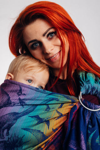 Model wearing baby in Lenny Lamb ring sling baby carrier in print Jurassic Park - New Era. Features Vibrant gradient background of purple to teal to yellow to red. Dark purple realistic-looking dinos and a on a dark purple weft. Close up